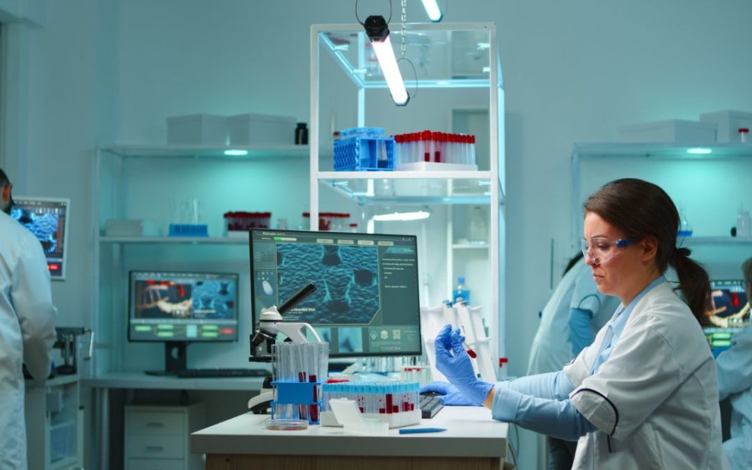 Investigadora en laboratorio analizando muestra de sangre frente a una computadora y microscopio.